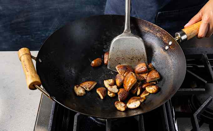 We see mushrooms cooking in a wok. A spoon/wok spatula is used to push it around.